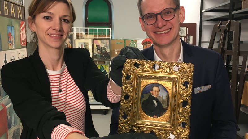 Zwei Museumsmitarbeiter halten ein historisches Portrait im Goldrahmen hoch.
