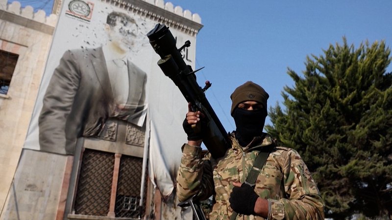 Ein vermummter und bewaffneter Kämpfer einer regierungsfeindlichen Miliz steht vor einem Gebäude, an der ein überlebensgroßes Plakat mit dem halb zerstörten Bild des Machthabers Assad hängt. In Syrien sind die regierungsfeindlichen Milizen offenbar weiter auf dem Vormarsch. Von Süden her rücken sie auf die Hauptstadt Damaskus vor. Es gibt Berichte, wonach die Regierungstruppen sich in mehreren Regionen weitgehend kampflos zurückziehen. | Bild: Bayerischer Rundfunk 2024 Ein vermummter und bewaffneter Kämpfer einer regierungsfeindlichen Miliz steht vor einem Gebäude, an der ein überlebensgroßes Plakat mit dem halb zerstörten Bild des Machthabers Assad hängt. In Syrien sind die regierungsfeindlichen Milizen offenbar weiter auf dem Vormarsch. Von Süden her rücken sie auf die Hauptstadt Damaskus vor. Es gibt Berichte, wonach die Regierungstruppen sich in mehreren Regionen weitgehend kampflos zurückziehen.
