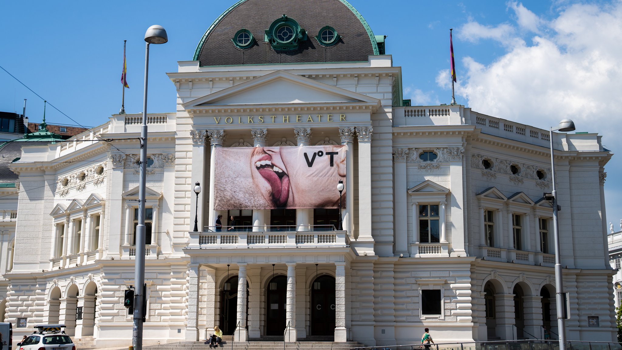 Bild des Wiener Volkstheaters, ein imposanter klassizistischer Bau, über dem Eingang hängt ein Transparent auf dem sich ein Mann und eine Frau leidenschaftlich küssen