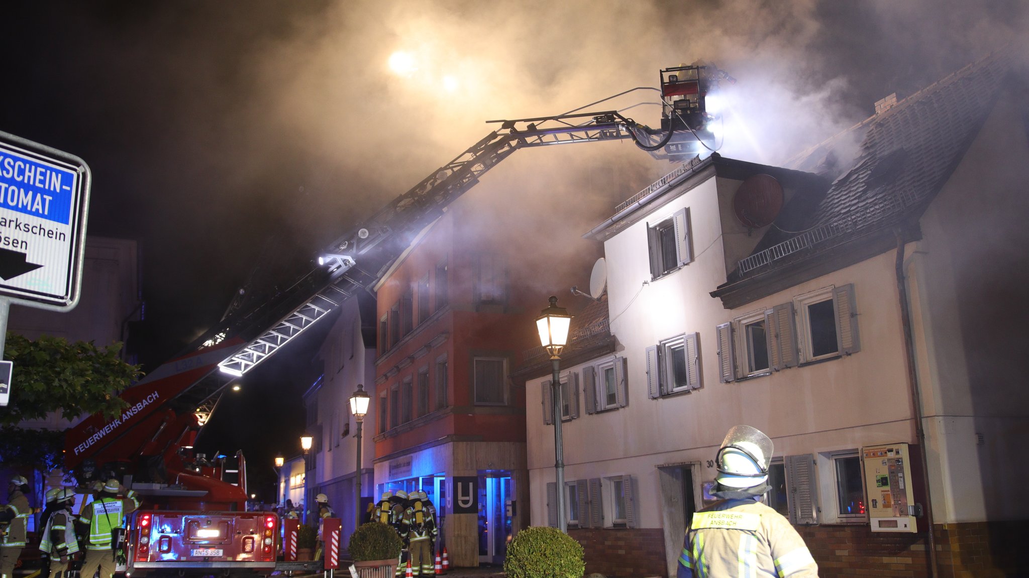 Einsatzkräfte der Feuerwehr stehen vor einem stark rauchenden Haus. Von einem Feuerwehrfahrzeug ist eine Leiter in Richtung des Gebäudes ausgerichtet. 