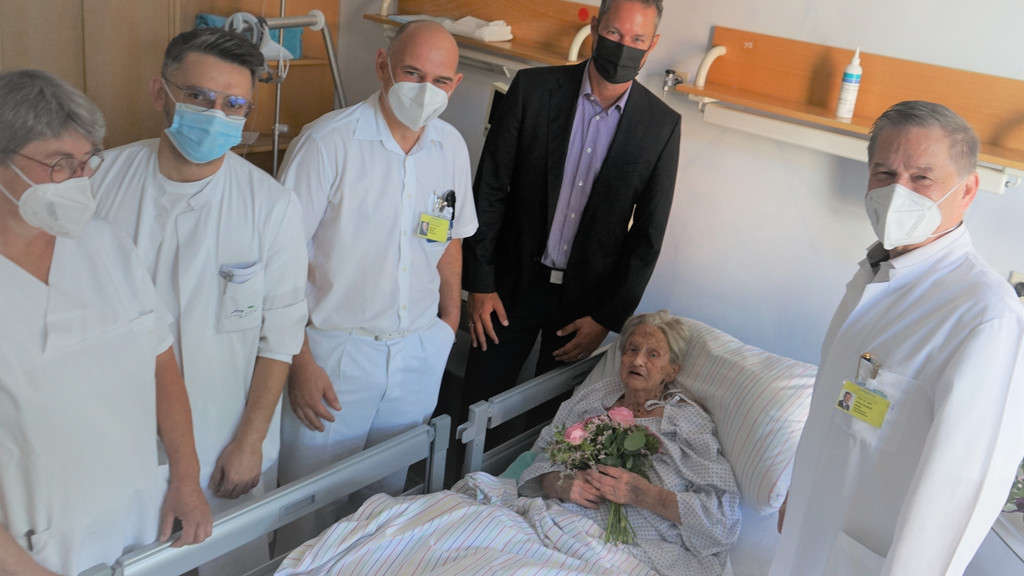 Eine Frau liegt mit einem Blumenstrauß im Krankenbett. Neben ihr stehen vier Männer und eine Frau mit Mund-Nasen-Schutz.