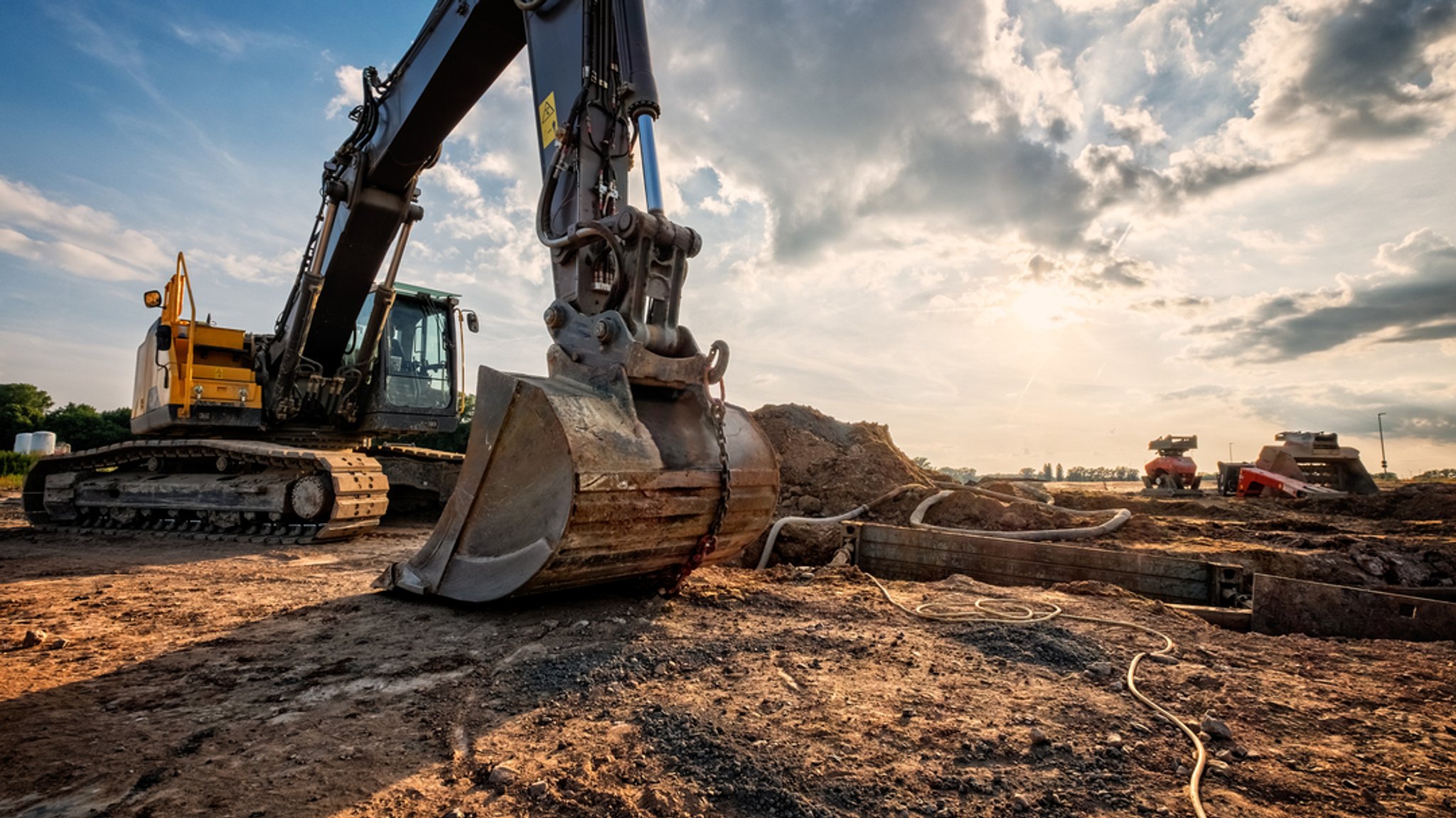(Symbolbild) Ein großer Bagger steht auf einer Baustelle im Erdreich bei Sonnenuntergang.