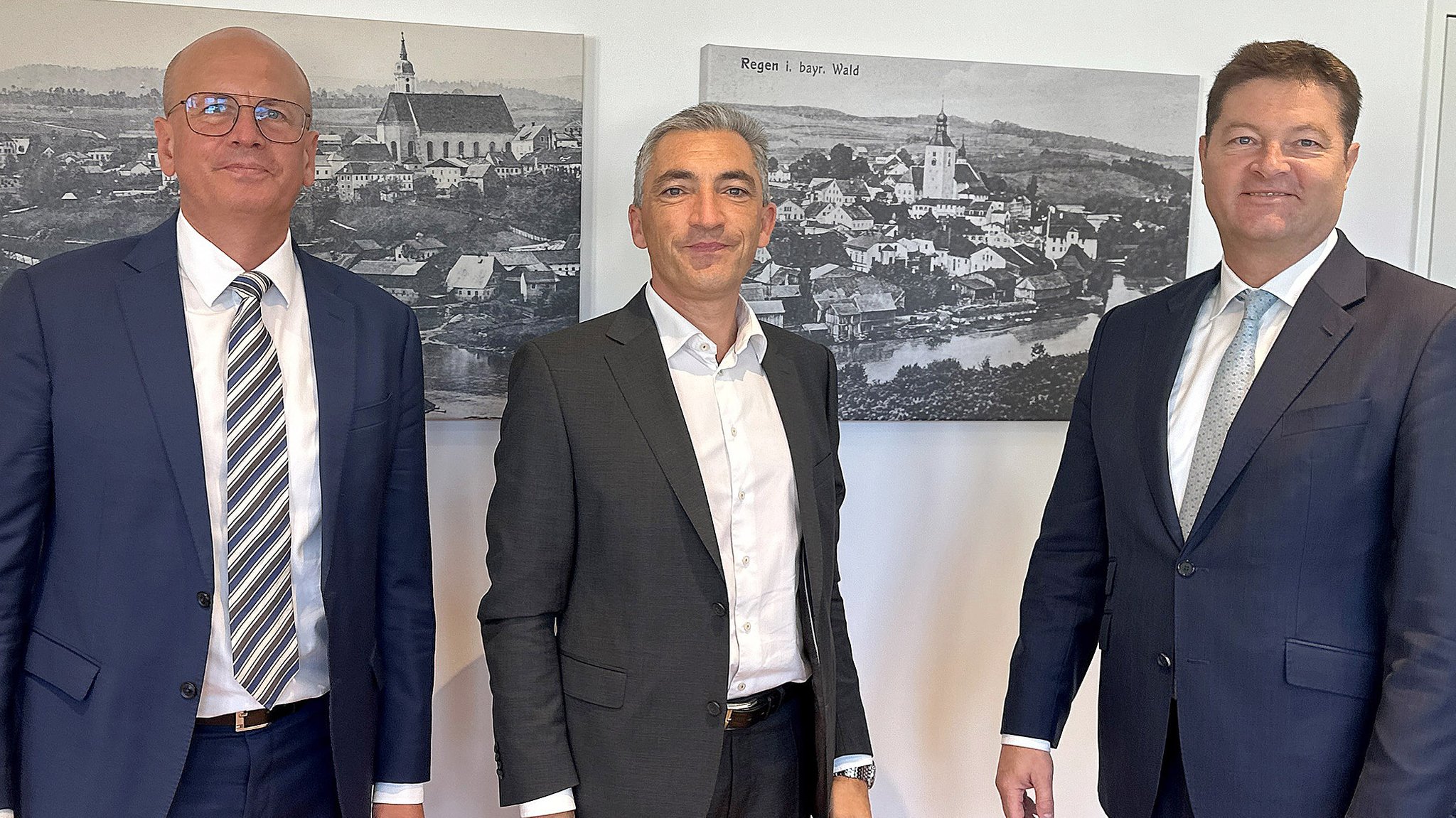 Landrat Ronny Raith (Bildmitte) mit Rodenstock CEO Marcus Desimoni (re.) und Rodenstock COO Roland Dimbarth stehen gemeinsam vor einer Wand mit Fotos.