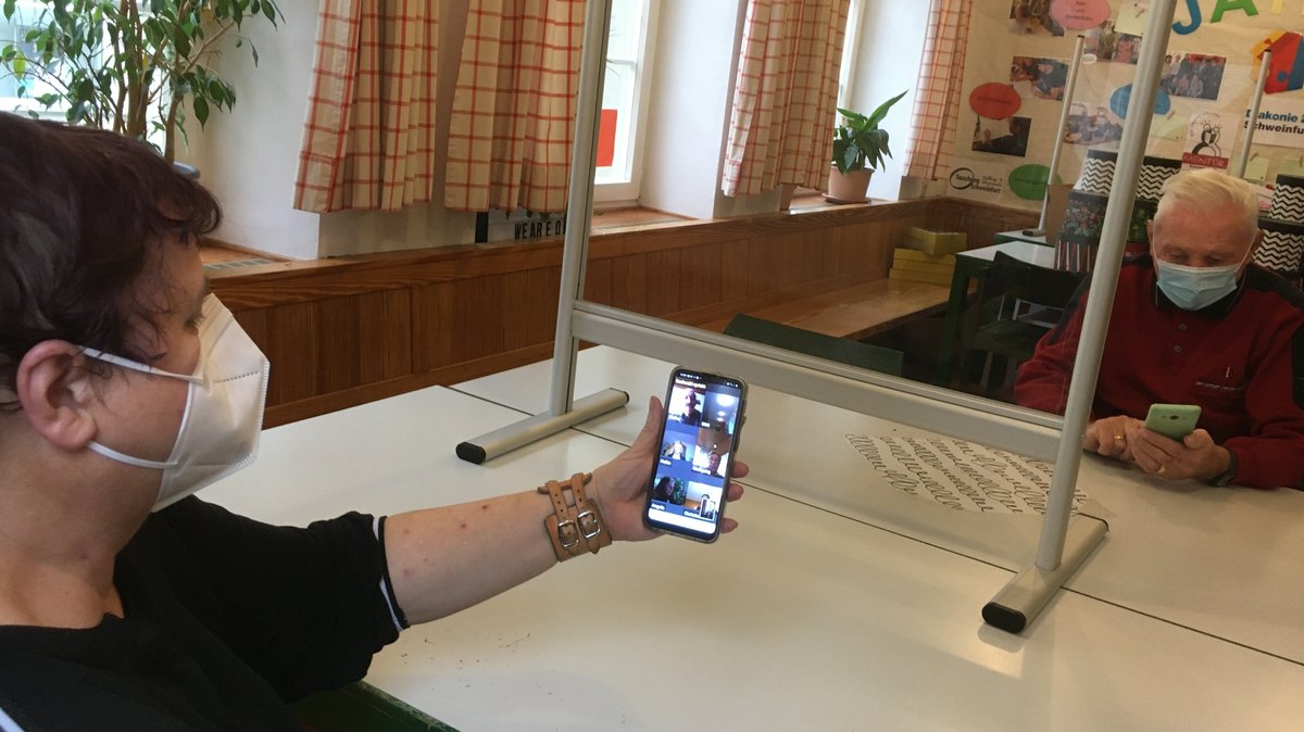 Ein Mitarbeiterin der Diakonie Schweinfurt erklärt einem Rentner, wie eine Videokonferenz auf dem Smartphone funktioniert.
