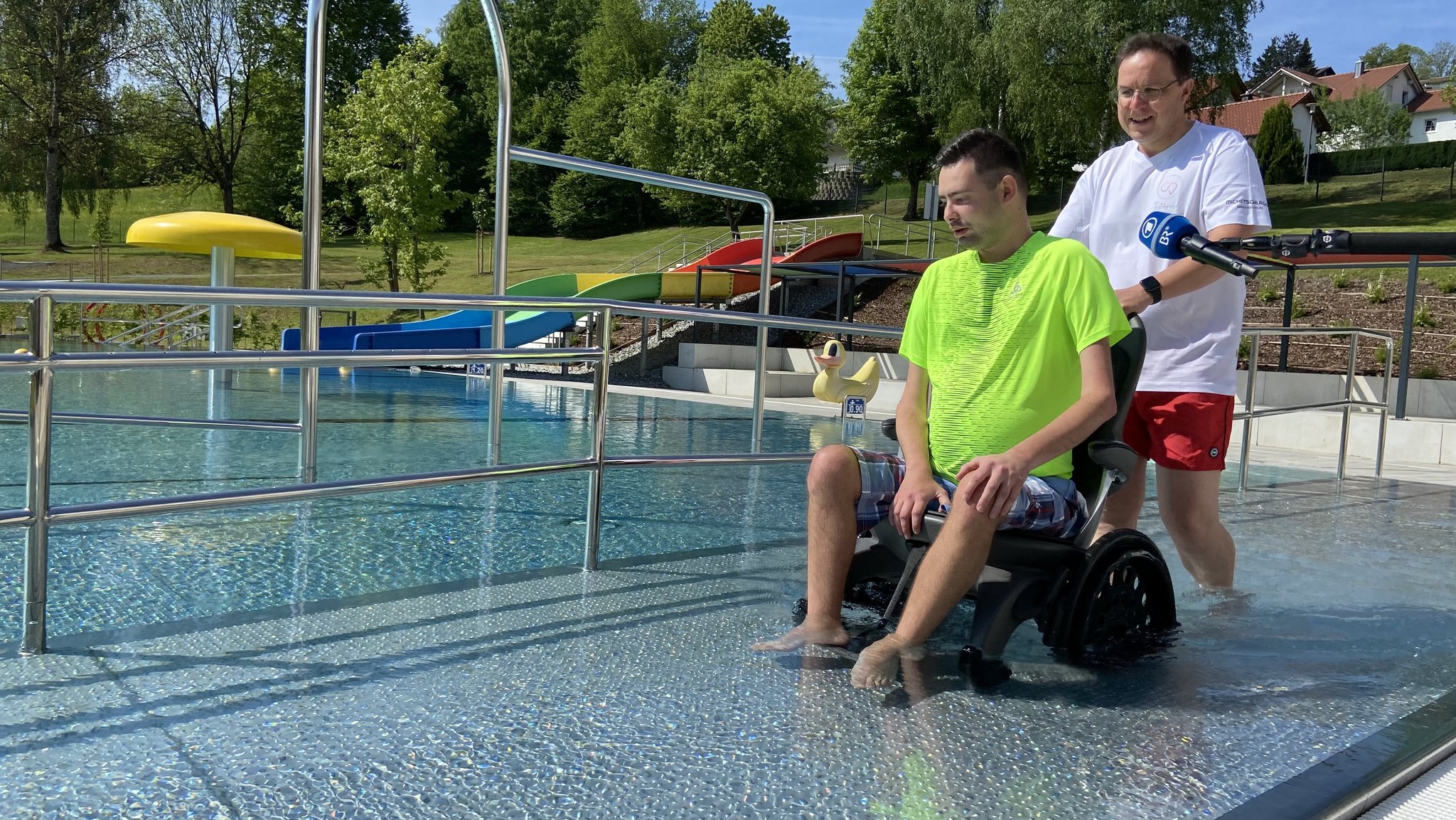 Ein junger Mann im Rollstuhl wird vom Bademeister ins barrierefreie Becken geschoben.