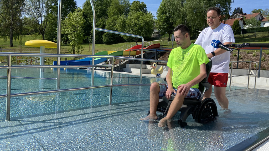 Ein junger Mann im Rollstuhl wird vom Bademeister ins barrierefreie Becken geschoben.