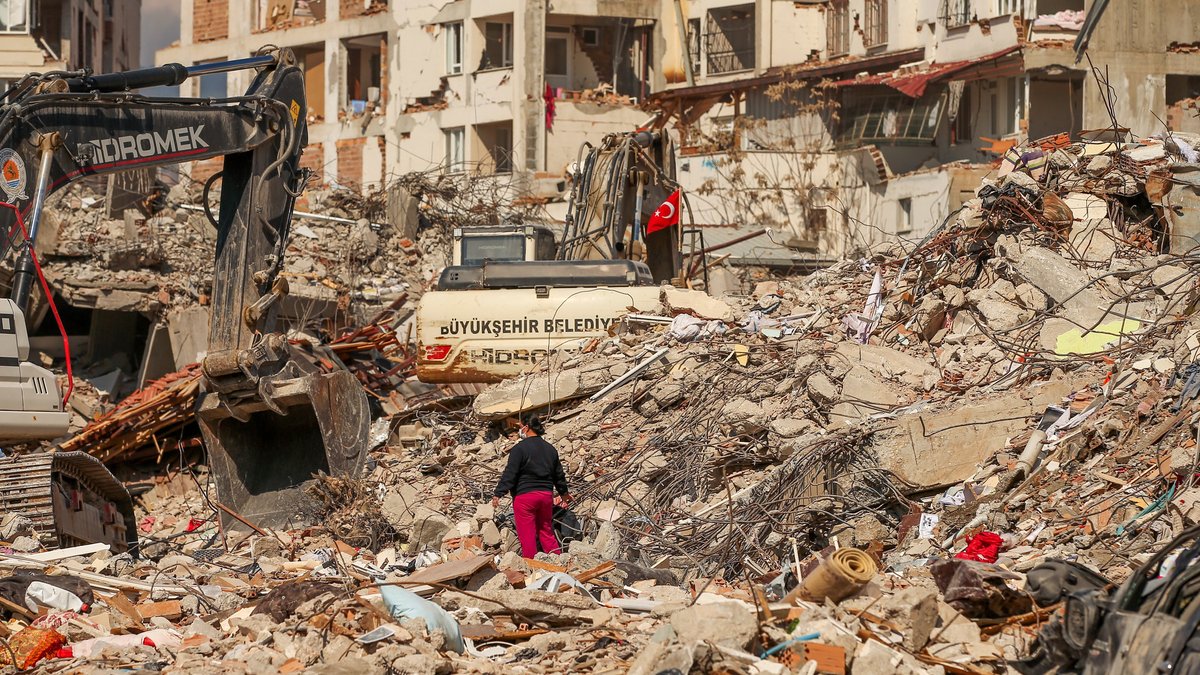 22.02.2023, Türkei, Samandag: Ein Mann steht zwischen Trümmern, während Bagger die Überreste eines zerstörten Gebäudes einreißen. 