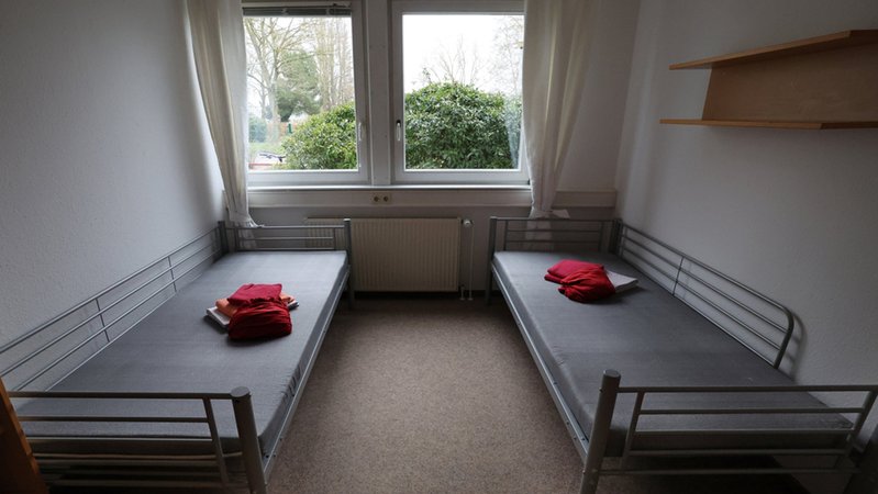 Ein Wohncontainer, in dem zwei leere, sehr einfache Betten stehen. Darauf liegt rote Bettwäsche. Am Ende des Raums sind zwei Fenster, vor denen Büsche stehen. | Bild: dpa-Bildfunk/Bernd Wüstneck Ein Wohncontainer, in dem zwei leere, sehr einfache Betten stehen. Darauf liegt rote Bettwäsche. Am Ende des Raums sind zwei Fenster, vor denen Büsche stehen.