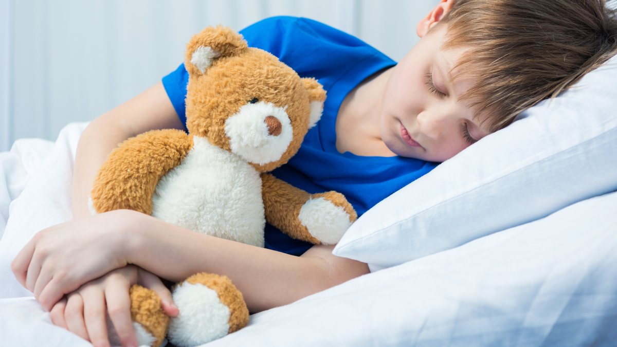 Krankes Kind im Bett mit Teddy-Bär im Arm.
