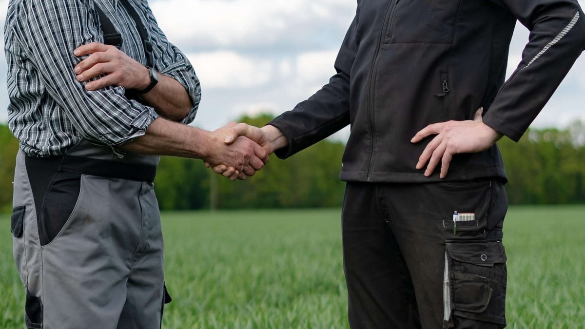Zwei Personen in Arbeitskleidung stehen auf einem Getreidefeld und reichen sich die Hand (Symbolbild)