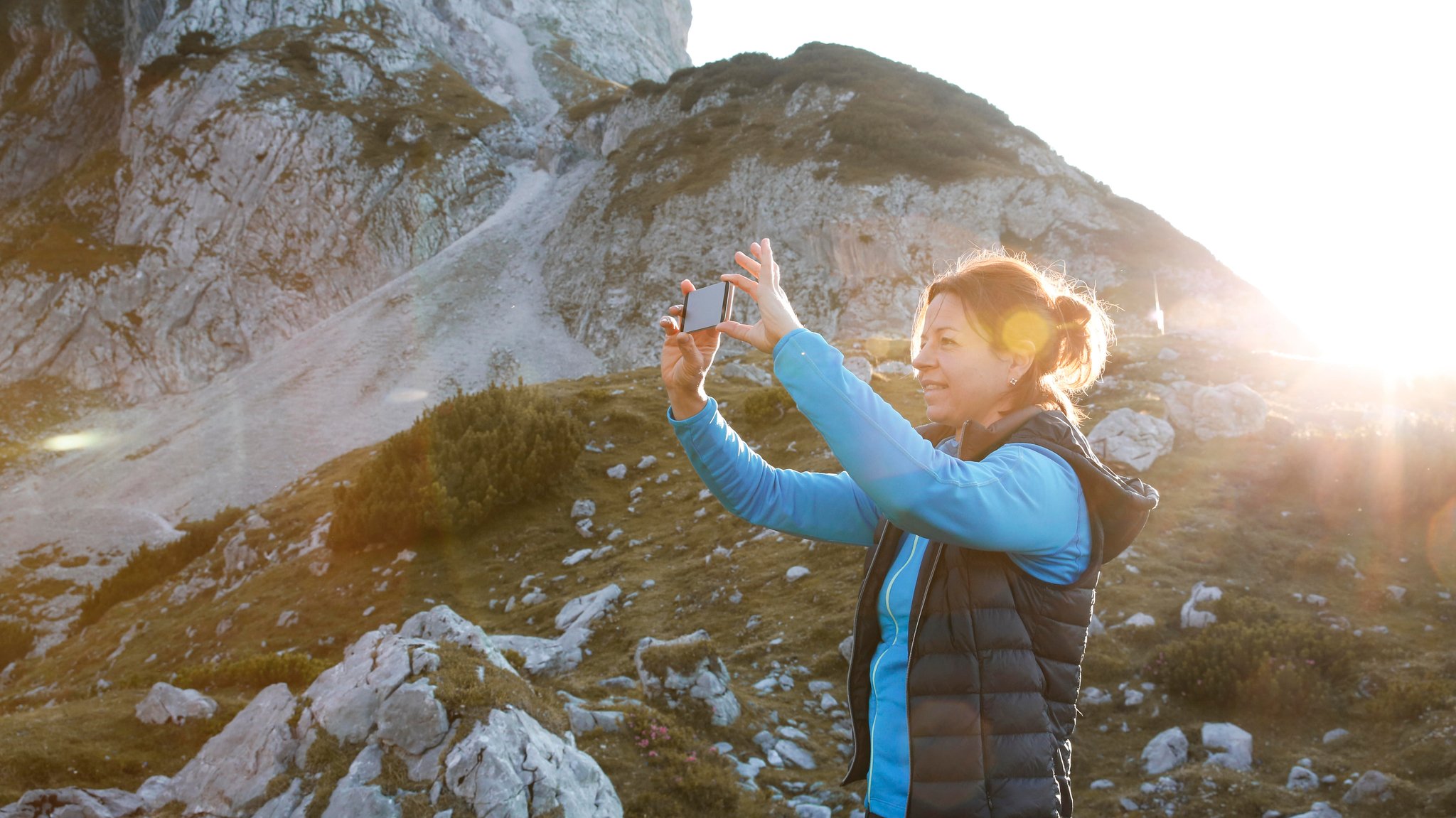 (Symbolbild) Eine Frau in Wanderklamotten macht mit ihrem Handy ein Foto, hinter ihr ist eine Bergkulisse erkennbar.