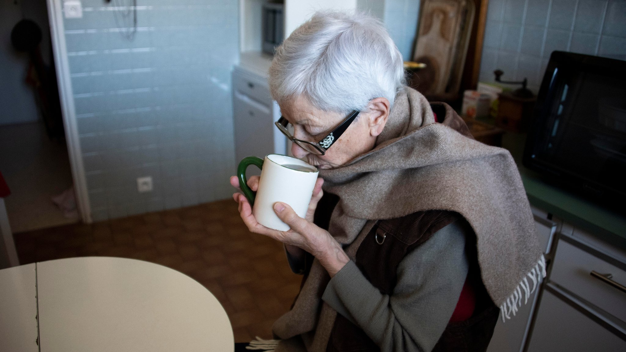 Ältere Frau sitzt mit Schaal und Teetasse am Küchentisch