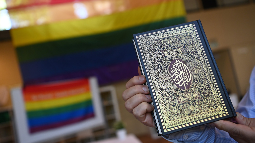 Ein Imam hält einen Koran vor eine Regenbogenflagge.