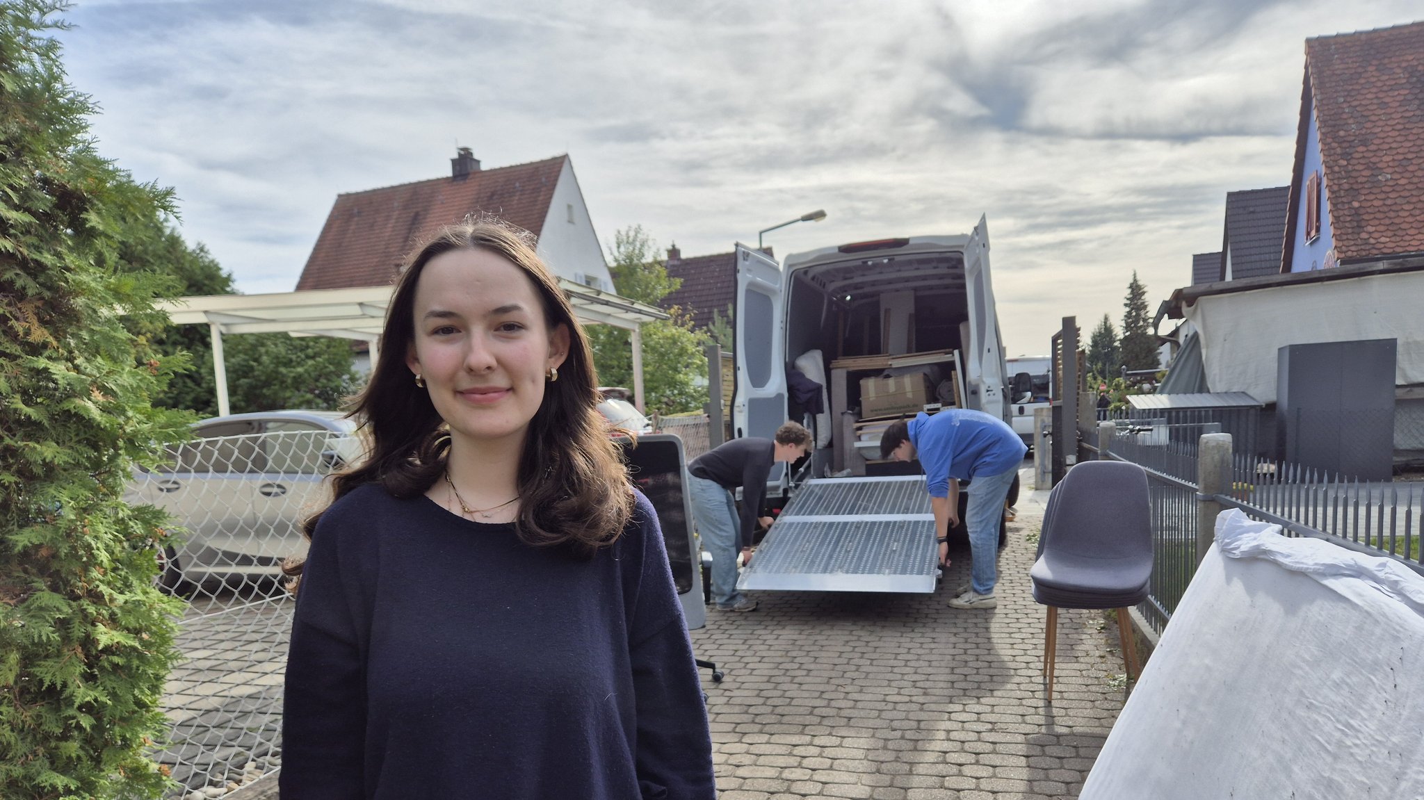 Lena steht in der Einfahrt ihres Elternhauses. Im Hintergrund beladen zwei Freunde den Umzugswagen. | Bild: Iris Tsakiridis / BR Lena steht in der Einfahrt ihres Elternhauses. Im Hintergrund beladen zwei Freunde den Umzugswagen.