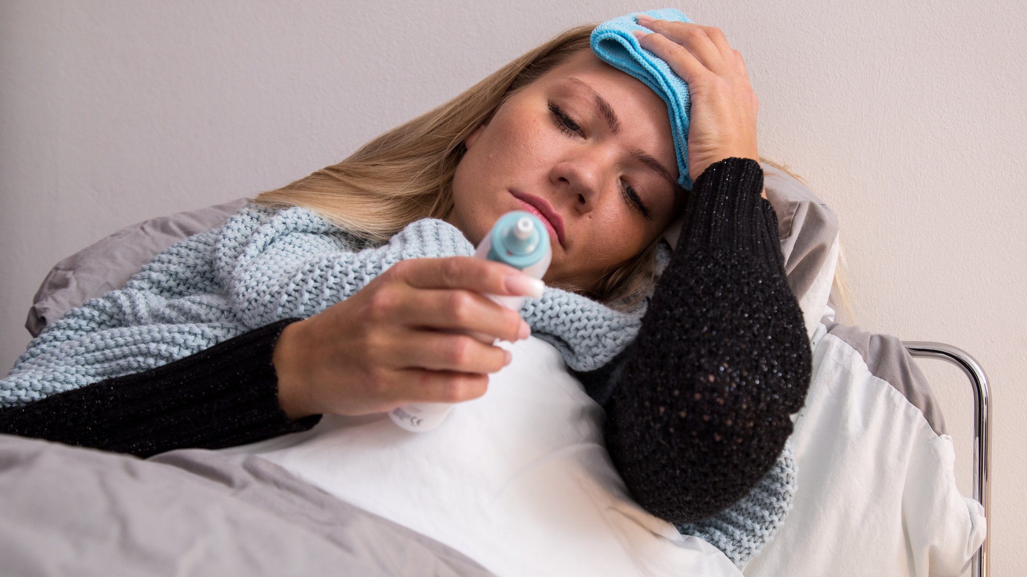 Symbolbild für Krankheit: Frau liegt mit Fieberthermometer im Bett und hält sich einen Lappen an die Stirn