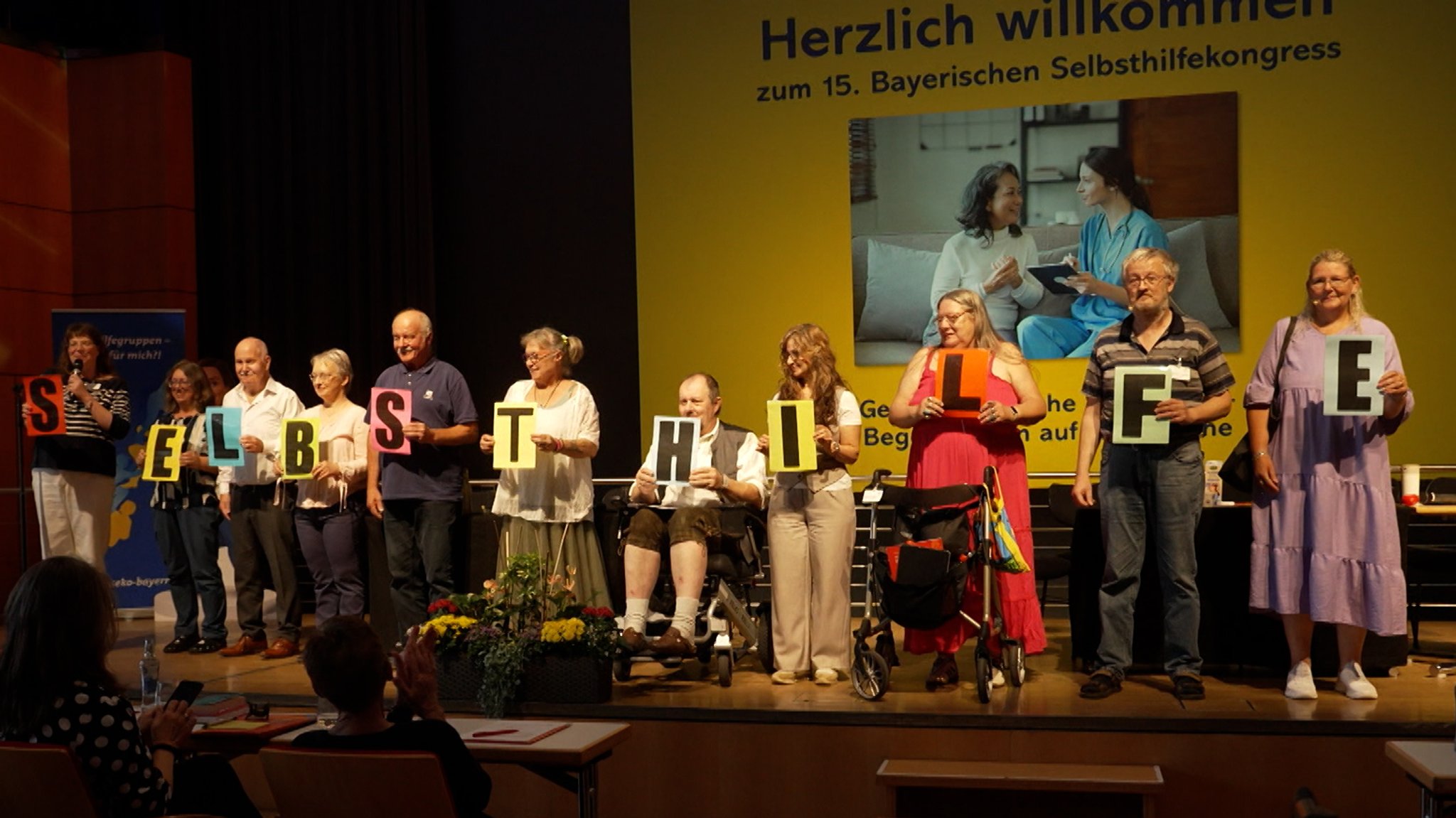 (Symbolbild) Mehrere Menschen auf einer Bühne halten in ihren Händen jeweils einen Buchstaben; zusammen ergibt es das Wort "Selbsthilfe". | Bild: Bernd Stuhlmüller/BR (Symbolbild) Mehrere Menschen auf einer Bühne halten in ihren Händen jeweils einen Buchstaben; zusammen ergibt es das Wort "Selbsthilfe".