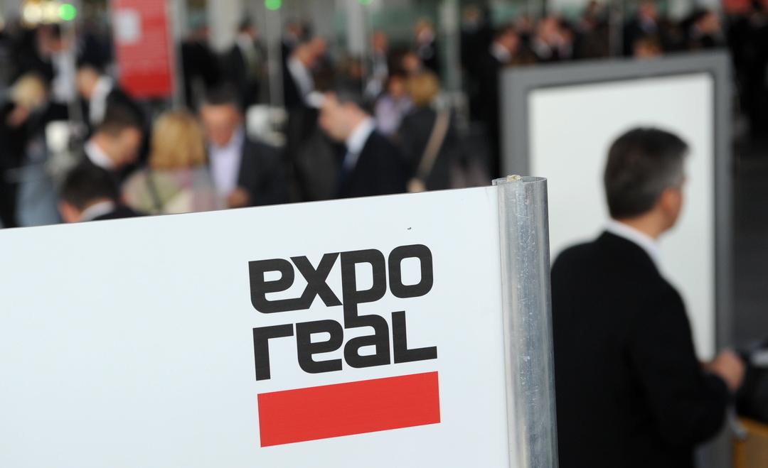 Besucher kommen zur Messe expo real und gehen an einem Logo der Messe vorbei.