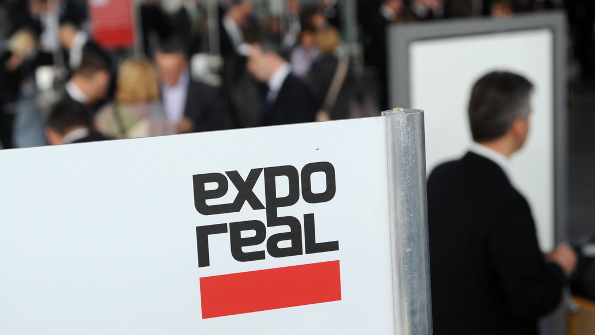 Besucher kommen zur Messe expo real und gehen an einem Logo der Messe vorbei.