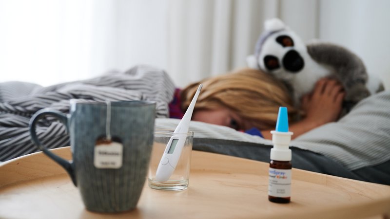 Fieberthermometer, Nasenspray und eine Tasse Tee stehen vor einem Bett, in dem ein Kind mit Kuscheltier liegt (Symbolbild).