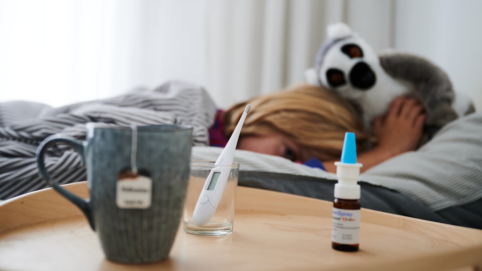 Fieberthermometer, Nasenspray und eine Tasse Tee stehen vor einem Bett, in dem ein Kind mit Kuscheltier liegt (Symbolbild).