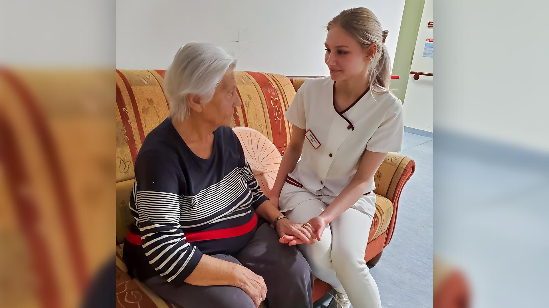 Pflegeschülerin Isabel sitzt auf einer Couch neben einer alten Frau und hält ihre Hand