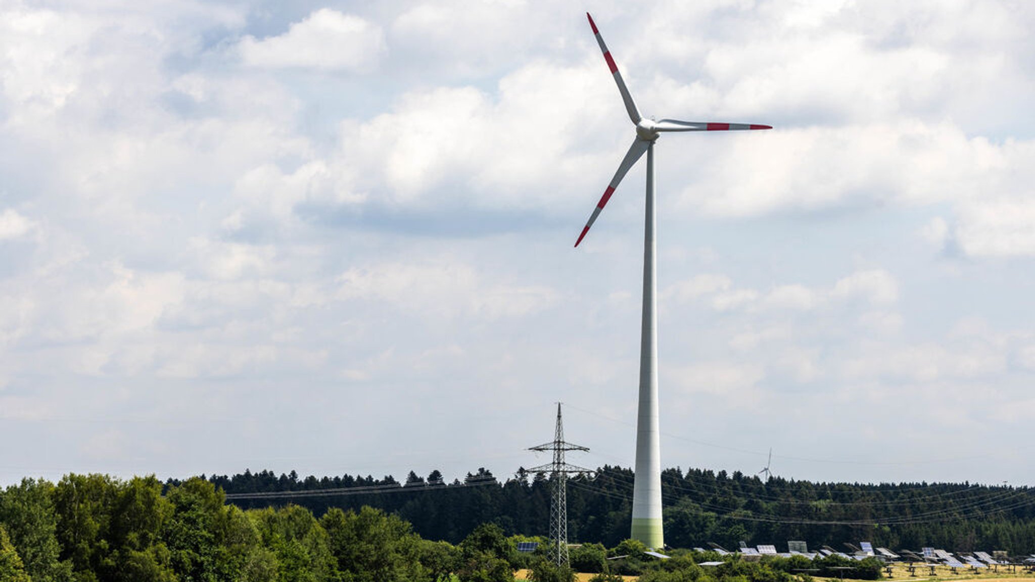 Ein Windrad steht in der Landschaft, während davor eine Hochspannungsleitung und Photovoltaikanlagen zu sehen sind. 