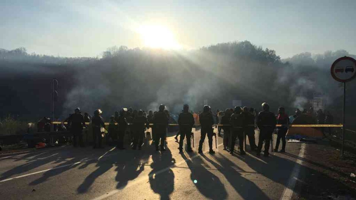 Polizisten mit Helmen und Schildern stehen am Grenzübergang einer Gruppe Menschen gegenüber. Einzelne Rauchschwaden ziehen auf.