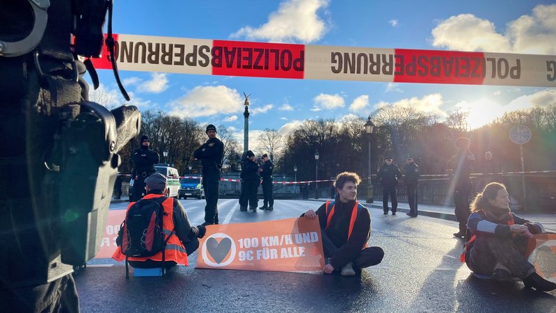 Klimaaktivisten sitzen auf der Straße und blockieren den Verkehr auf der Münchner Luitpoldbrücke. Der Bereich ist von der Polizei abgesperrt.