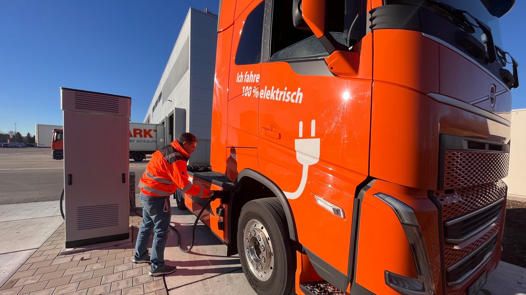 Ein Mann in orange-farbener Weste verbindet einen orange-farbenen LKW mittels Stecker mit einer Ladesäule.