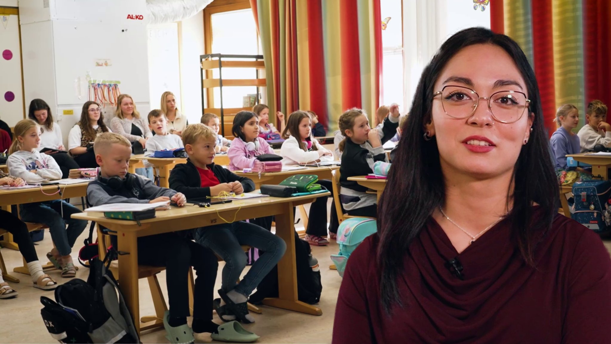 Ebru im Portrait vor einem Klassenraum mit Kindern. Sie ist Lehramtsanwärterin im Vorbereitungsdienst an der Grundschule Burgkirchen an der Alz. Sie unterrichtet Englisch im Hauptfach und Deutsch, Mathematik und Kunst im Nebenfach. 