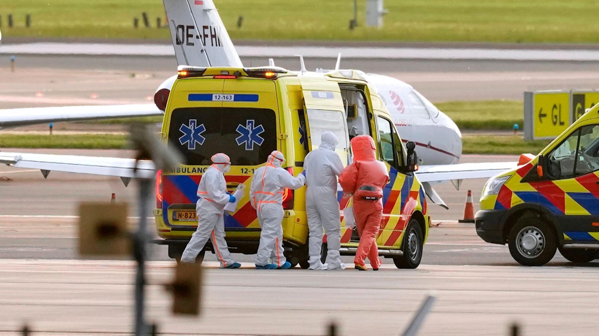 Personen in Schutzanzügen stehen bei einem niederländischen Krankenwagen auf einem Rollfeld