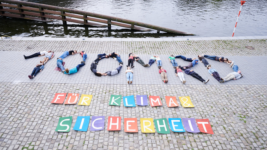 10 MRD für Klima & Sicherheit steht auf einem Boden geschrieben. Die Buchstaben und Zahlen bei "100 MRD" sind durch am Boden liegende Menschen dargestellt. 