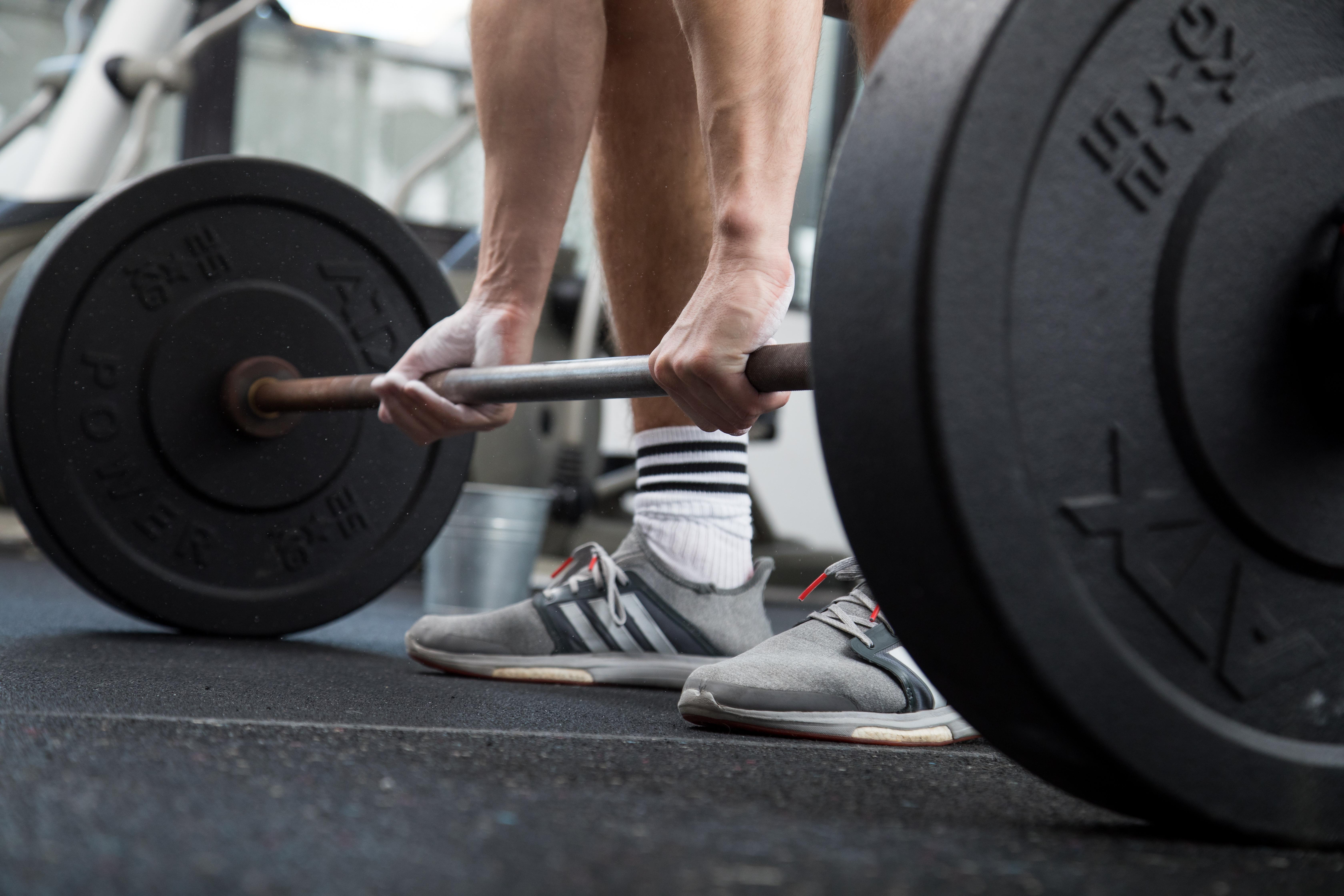 Symbolbild Gewichtheben: Zu sehen sind zwei Füße und zwei Hände, die nach einer Stange mit zwei schweren Gewichten greift
