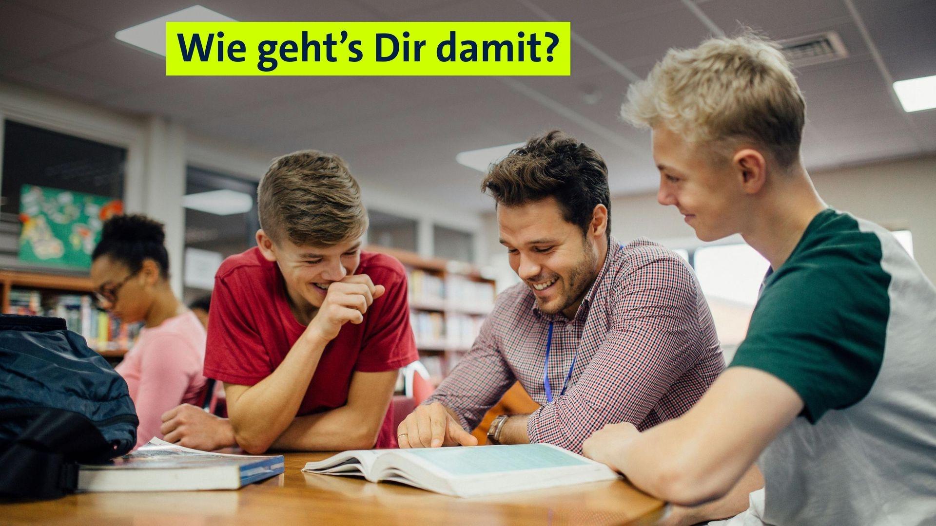 (Symbolbild) Ein Lehrer liest mit zwei Schülern in einem Schulbuch