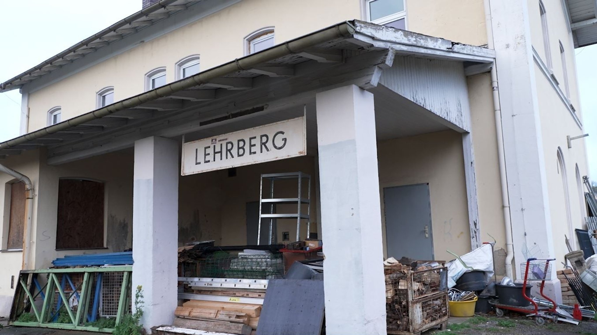 Bahnhofsschild mit der Aufschrift "Lehrberg" an einem alten Bahnhofsgebäude mit Müll drumherum