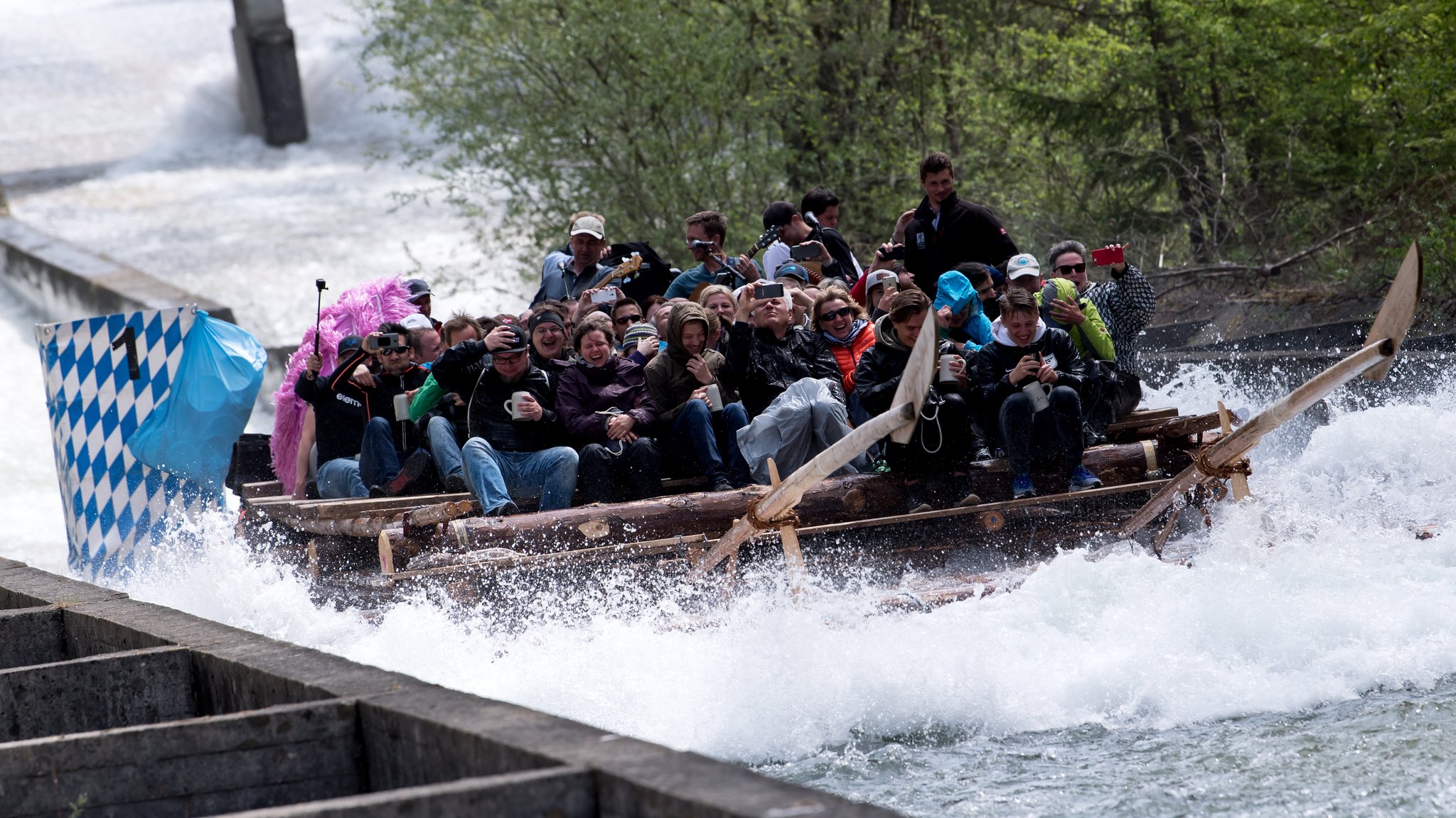 Viele Menschen auf einem Floß; sie rutschen eine Floßrutsche hinunter. Das Wasser spritzt, die Menschen lachen und halten ihre Handys, Bierkrüge und Mützen fest.