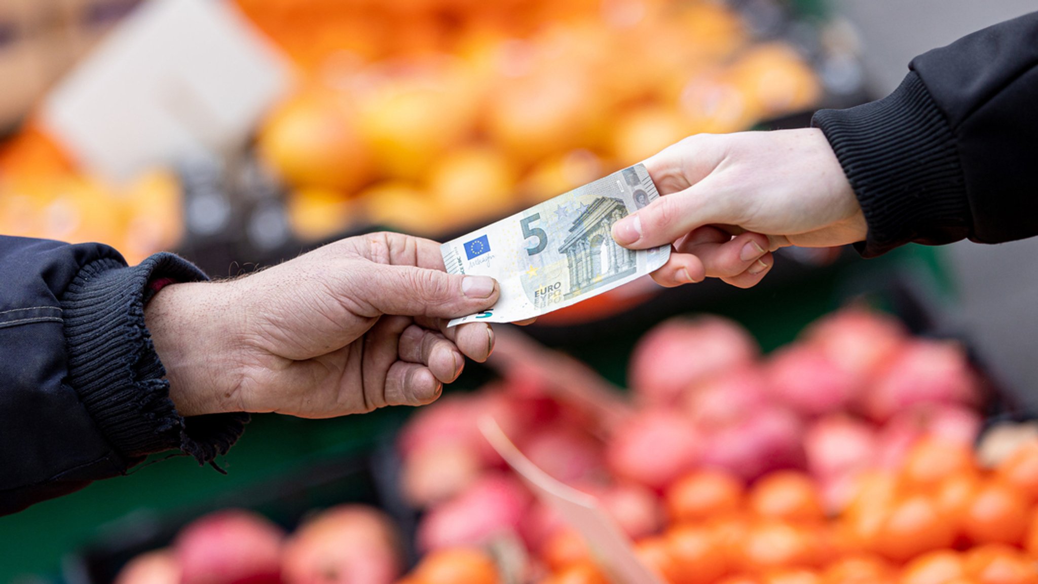 Ein Kunde bezahlt seinen Einkauf an einem Stand auf einem Wochenmarkt mit einem Fünf-Euro-Schein.