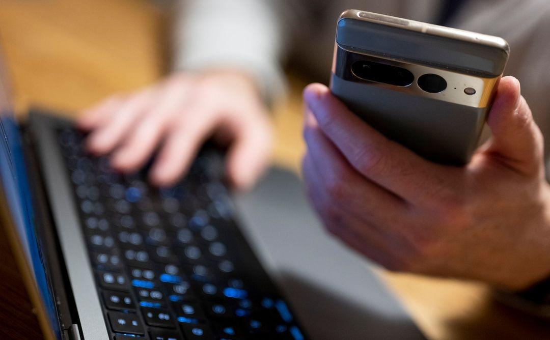 Ein Mann sitzt am Laptop und hält ein Smartphone in der Hand.