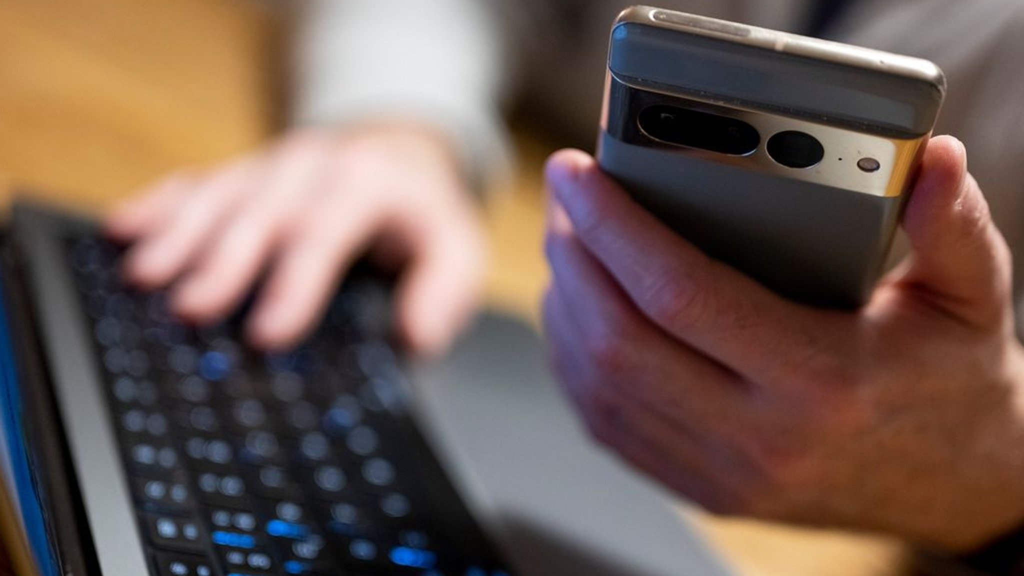 Ein Mann sitzt am Laptop und hält ein Smartphone in der Hand.