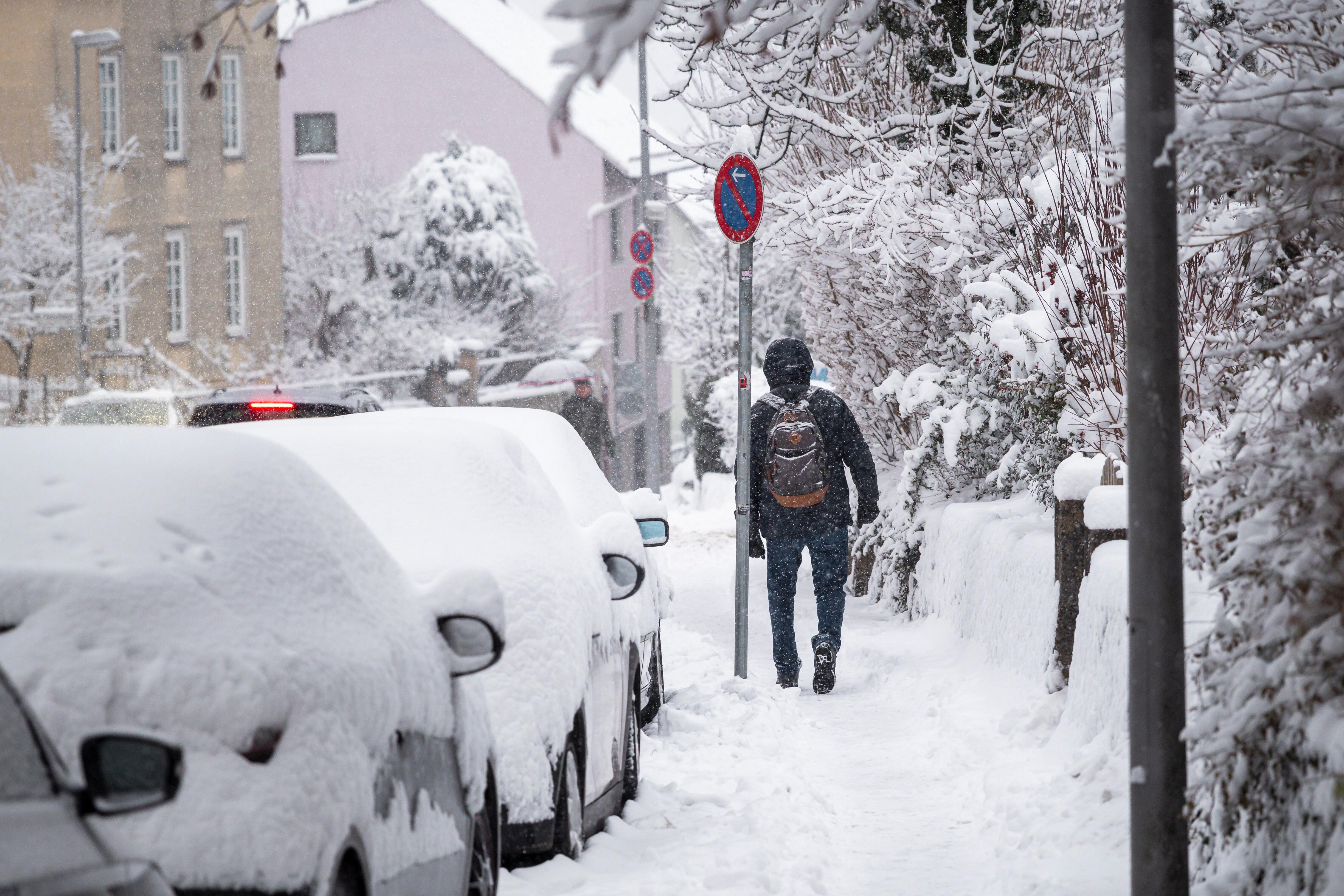 Eine Person geht auf einem verschneiten Gehweg entlang. Die parkenden Autos sind mit Schnee bedeckt. Im Verlauf des Tages gibt es weitere Schneefälle und Glätte. (26.01.2026)