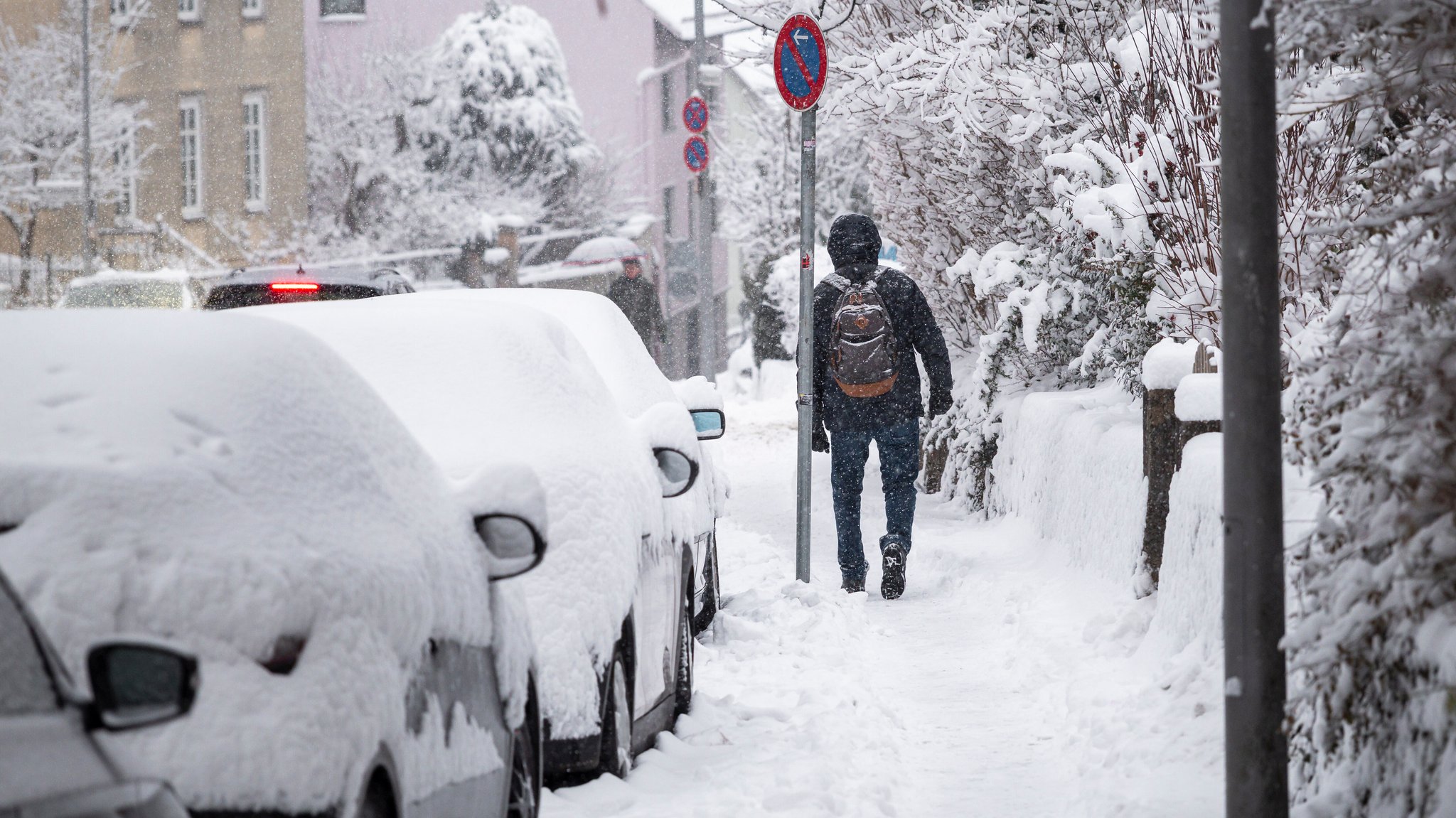 Eine Person geht auf einem verschneiten Gehweg entlang. Die parkenden Autos sind mit Schnee bedeckt. Im Verlauf des Tages gibt es weitere Schneefälle und Glätte. (26.01.2026)