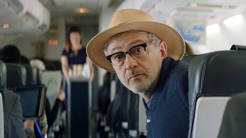 Elia Suleiman blickt in "Vom Gießen des Zitronenbaums", aus einem Flugzeugsitz gelehnt, in die Kamera (Filmszene)