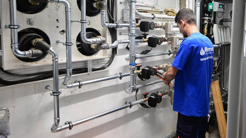 Ein Mitarbeiter in einem Handwerksbetrieb für Heizungsbau, arbeitet in einem Heizungskeller einem Sportheim an der Installation der Technik für eine Wärmepumpe.