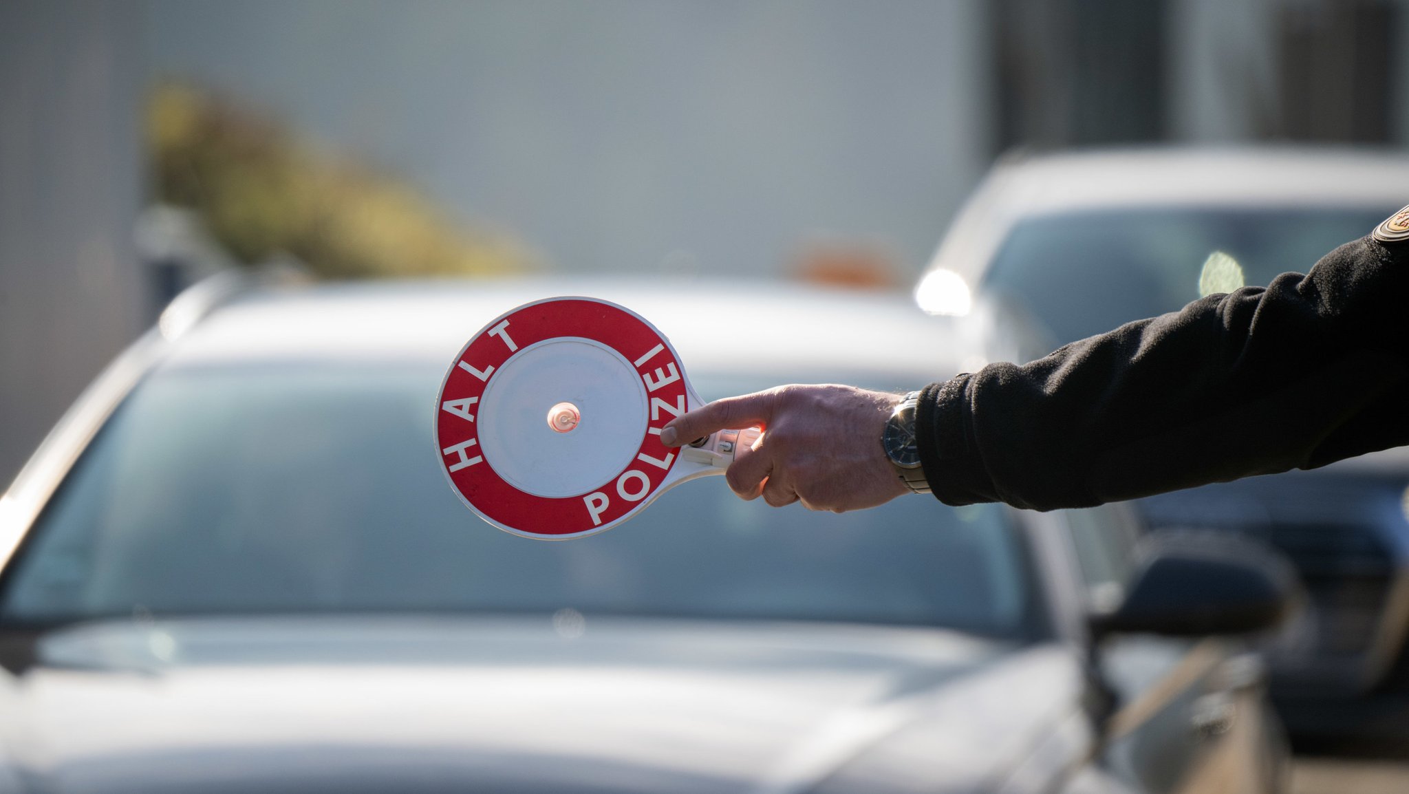 (Symbolbild) Eine Hand hält ein rundes, rot-weißes Schild, auf dem steht: "HALT POLIZEI". Dahinter sieht man zwei Autos hintereinander auf einer Straße.