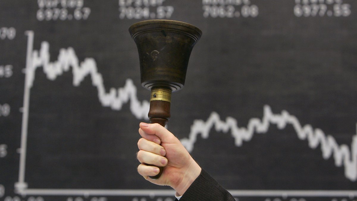 Symbolbild: Ein Mann hält eine Glocke vor der Anzeigentafel des Deutschen Aktien Index (DAX).