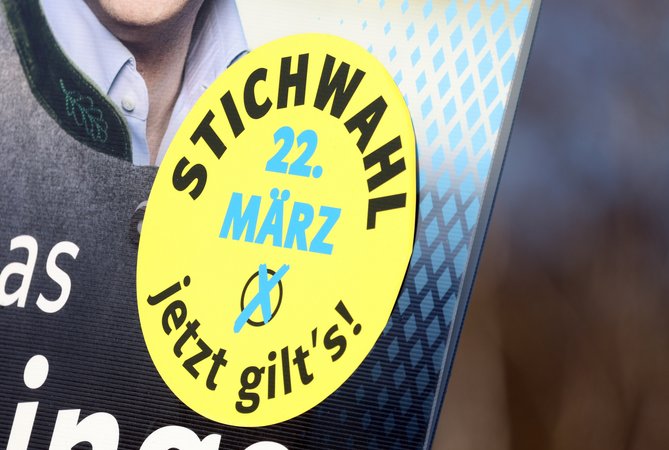 Auf einem Wahlplakat ist eine Plakette mit der Aufschrift "Stichwahl 22. März jetzt gilt's" aufgeklebt.