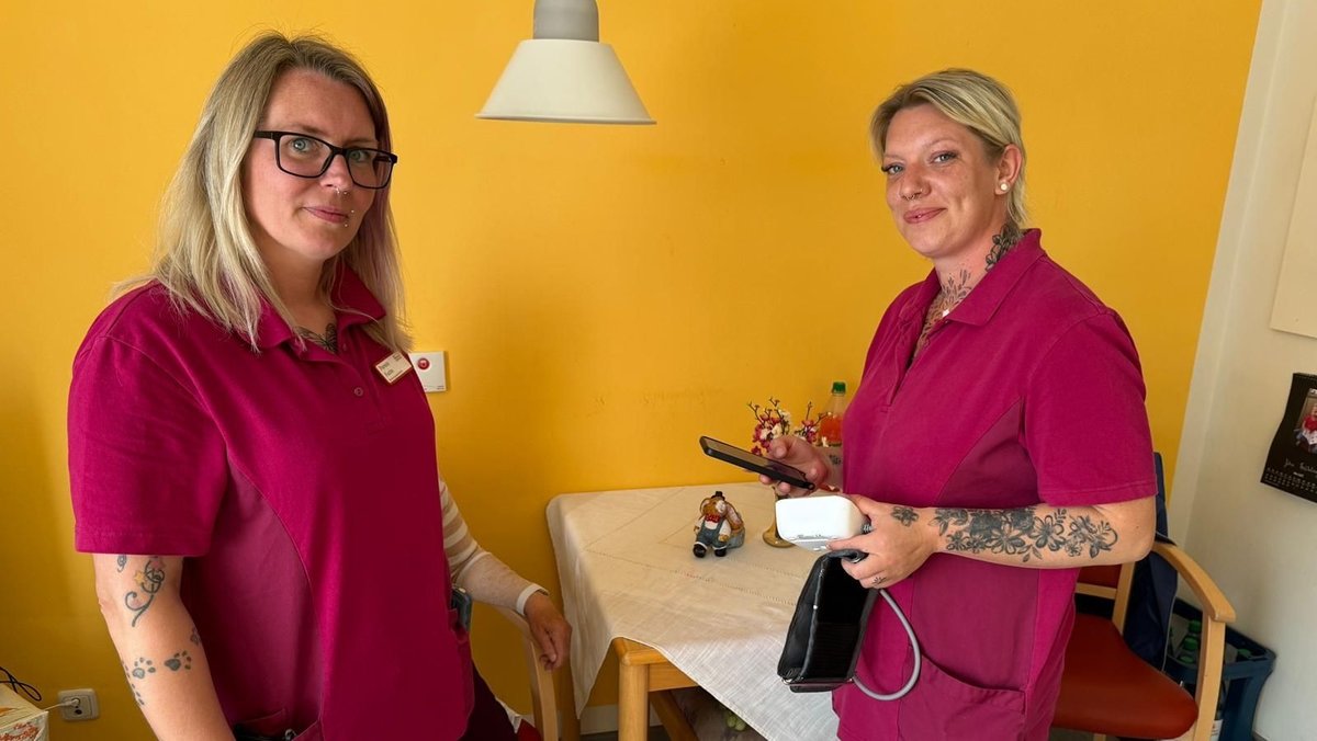 Zwei Pflegerinnen in bordeauxroter Arbeitskleidung stehen vor einer gelben Wand. Eine von ihnen hält ein Blutdruckmessgerät und ein Smartphone in der Hand.