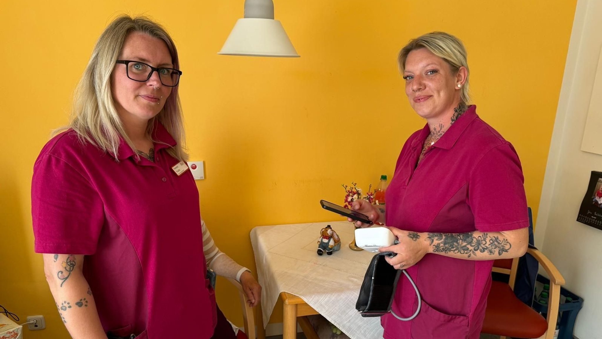Zwei Pflegerinnen in bordeauxroter Arbeitskleidung stehen vor einer gelben Wand. Eine von ihnen hält ein Blutdruckmessgerät und ein Smartphone in der Hand. | Bild: Diakonie Hochfranken Zwei Pflegerinnen in bordeauxroter Arbeitskleidung stehen vor einer gelben Wand. Eine von ihnen hält ein Blutdruckmessgerät und ein Smartphone in der Hand.