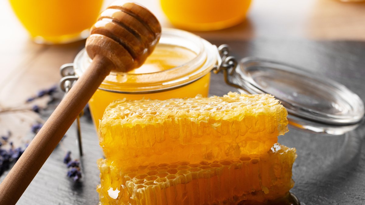 Honiglöffel über einem Glas Honig, davor zwei Honigwaben.