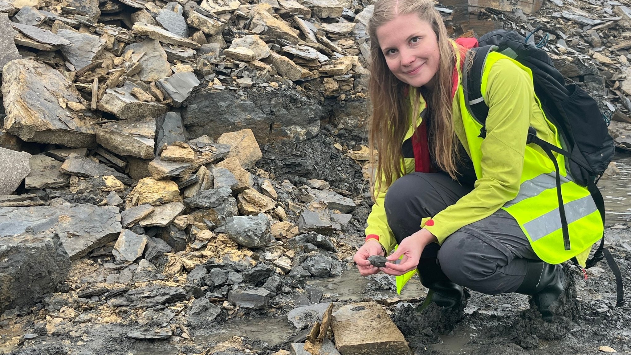 Eine Frau kniet vor Gestein und hält einen Brocken in der Hand. 